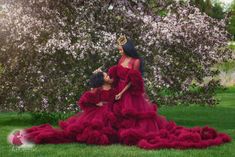 two women in red gowns sitting on the grass with trees and flowers behind them