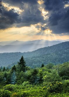 the sun shines through clouds over mountains and trees in the foreground, as seen from an overlook