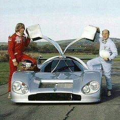 two men standing next to a silver sports car