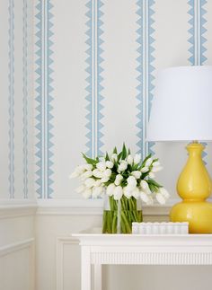 a yellow vase with white flowers on a table next to a lamp and wallpaper