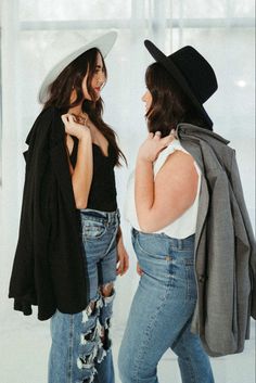 two women in jeans and hats standing next to each other with jackets on their shoulders