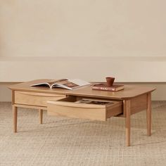 a coffee table with an open book on top and two drawers underneath it, in front of a beige wall