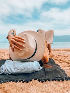 a person laying on top of a beach next to the ocean wearing a hat and reading a book