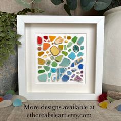 a white frame holding a multicolored sea glass mosaic in front of a potted plant