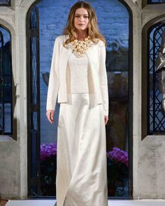 a woman is walking down the runway in a white dress and jacket with gold accents