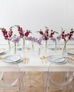 the table is set with clear chairs, plates and vases filled with purple flowers