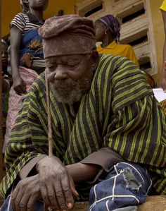 an old man sitting on the ground with his hands in his pockets and other people standing around him