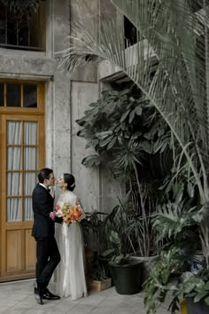 a bride and groom standing in front of a building with plants on the side walk