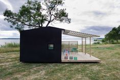 a small black house with a wooden deck and pergolated roof in the middle of an open field