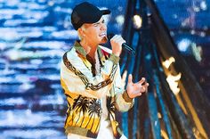 a woman singing into a microphone in front of a large screen with buildings behind her