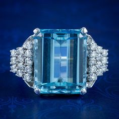 A magnificent vintage cocktail ring made in the Art Deco fashion with a colossal emerald cut aquamarine in the centre, weighing approx. 15ct with a pleasing, deep ocean blue hue.  The aqua is complemented by an audience of twenty-four sparkling brilliant cut diamonds arranged in a graduated, stepped design down each shoulder. They're 0.05ct each (approx. 1.20ct total) and have superb SI1 clarity - H colour.  The robust gallery is modelled entirely in 18ct white gold and displays the stones high Formal Light Blue Aquamarine Diamond Ring, Formal Aquamarine Diamond Ring With Brilliant Cut, Aquamarine Brilliant Cut Diamond Ring For Formal Occasions, Elegant Emerald Cut Topaz Ring With 17 Jewels, Luxury Blue Baguette Cut Topaz Ring, Elegant Emerald Cut Topaz Ring, Elegant Light Blue Aquamarine Diamond Ring, Luxury Blue Topaz Ring Gia Certified, Blue Gia Certified Emerald Ring For Formal Occasions