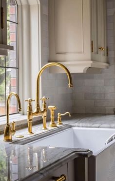 a kitchen sink with gold faucets and marble counter tops in front of two windows