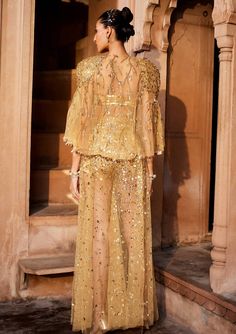 A bold three piece dress in sheer fabric, a glamorous embellished gold bralet paired with sheer trousers with splatter embroidery of sitara/patra and a matching sheer jacket with power shoulder highlighted with gold embellishments Party Pant Set With Sheer Dupatta, Festive Reception Dress With Sheer Sleeves, Evening Sets With Mirror Work In Organza, Festive Palazzo Set With Sequins And Cape Sleeves, Evening Palazzo Set With Sheer Dupatta For Eid, Evening Palazzo Set With Sheer Dupatta, Sequined Palazzo Set With Cape Sleeves For Parties, Glamorous Sequined Sets For Eid, Glamorous Sequin Sets For Eid