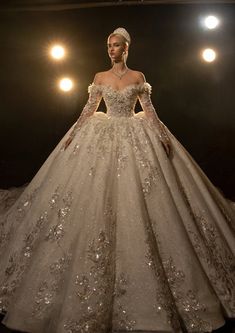 a woman in a wedding dress with long sleeves and flowers on the skirt is standing in front of spotlights