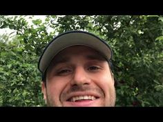 a close up of a person wearing a baseball cap and smiling at the camera with trees in the background