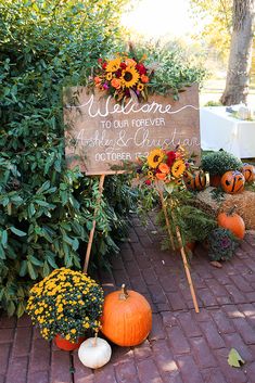 a welcome sign surrounded by pumpkins and flowers on the side of a brick walkway