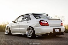 the back end of a white car parked on top of a parking lot next to tall grass