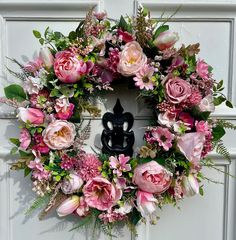 a wreath with pink flowers and greenery hanging on a white front door, next to a black statue