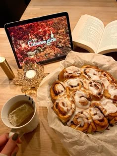 a person holding a cup of coffee next to a plate of cinnamon rolls and an open book