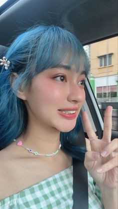 a woman with blue hair is sitting in a car and making the peace sign while wearing a green gingham top