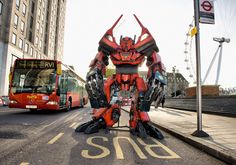 a giant robot standing on the side of a road next to a red bus and buildings