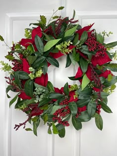 a wreath with red flowers and greenery hanging on a white front door, surrounded by green leaves