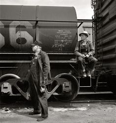 two men standing next to each other near a train on the tracks and one is looking at another man