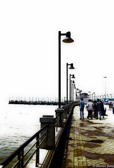 people are walking on the pier near the water and light poles with street lamps in front of them