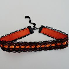 "This choker style necklace is made in layers of black lace stitched with an orange glitter ribbon. The lace and ribbon are crimped with black bars and linked to a matching extension chain. There is a black bead dangle linked on the chain. The choker closes on the first link at 13 \" and with the black extension chain adds adjustable sizing to 15\".  There is a matching lobster claw closure on the choker." Halloween Party Choker Jewelry, Edgy Halloween Party Choker, Halloween Black Chain Choker, Adjustable Black Halloween Choker, Edgy Halloween Choker Necklace, Ribbon Choker Necklace, Ribbon Choker, Choker Style Necklace, Black Bar