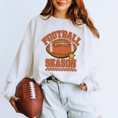 a woman holding a football in her right hand and wearing a sweatshirt that says football season on it