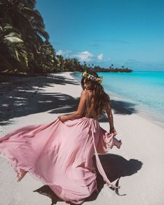 a woman in a pink dress is walking on the beach with palm trees behind her