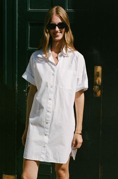 a woman standing in front of a green door wearing sunglasses and a white shirt dress