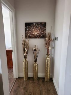 three gold vases with dried flowers in them on a wooden floor next to a painting