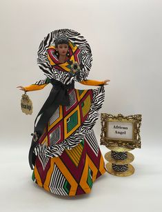 "This exquisite African Queen Figurine is a stunning blend of African Gold and multi colored diamond print fabric with a contrasting zebra print fabric that adds an ethnic and festive twist to its unique design.  Let me tell you about this African Queen: * She is a 17 Inch Figurine, made with a vinyl head and hands with bendable arms, affixed to an open bottom cone. *  Her long sleeve dress is made in large African print diamond pattern fabric with tunic underlay, her overskirt is made in smaller print fabric in an asymmetrical design to create the illusion of movement, trimmed in ruffled zebra trim and tied around her waist is a black satin ribbon bow tied in the front.  The look is completed with a breathtaking matching high collar caplet and black pearled and gold pendants, as well as l African American Christmas Figurines, African American Tree Toppers, African American Angel Tree Topper, African Christmas Tree Topper, African Crafts Walmart, African American Christmas Walmart, Afro American Figurines, African American Art Dolls, African American Themed Christmas Tree