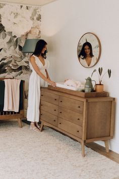 a woman standing next to a dresser with a baby in it and a mirror on the wall