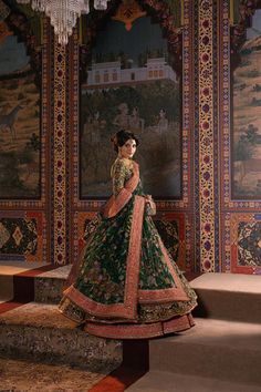 a woman in a green and gold wedding dress standing on stairs with an elaborate painting behind her