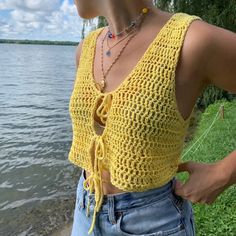 a woman standing next to the water wearing a yellow crochet top and jeans