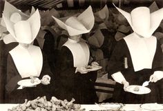 black and white photograph of people dressed in hats with plates of food on table next to them