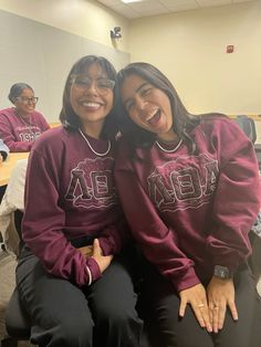 two women sitting next to each other in front of desks wearing sweatshirts with letters on them