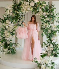 a woman in a pink dress standing next to a flower arch with white flowers and greenery