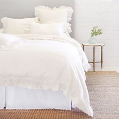 a white bed with ruffles and pillows in a room next to a table