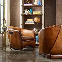 two brown leather chairs sitting next to each other in front of a bookshelf