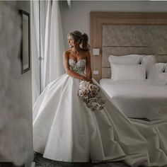 a woman in a wedding dress is standing near a bed and looking out the window