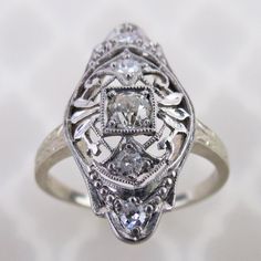 An old European cut diamond is centered on this antique 14k white gold filigree ring. The 3mm .10 carat precious gem is mounted in a square setting. Delicate airy scroll filigree with leaves and millgrain dots decorates the ring. The 13/16 inch long face has a domed gallery with engraved profiles, which is emulated on the ring's shoulders. There are four additional 1.5mm to 1.8mm diamonds set around on the ring. Collectively, this ring has an estimated total weight of .18 cwt diamonds.  The insi Antique Engraved White Gold Ring With Intricate Design, Antique White Gold Engraved Ring With Intricate Design, Vintage Diamond Jewelry With Intricate Design, Ornate White Gold Jewelry With Rose Cut Diamonds, Silver Jewelry With Rose Cut Diamonds For Vintage Events, Antique 14k White Gold Jewelry With Rose Cut Diamonds, Antique Diamond Ring With Rose Cut For Anniversary, Antique Diamond Ring With Rose Cut Diamonds For Anniversary, Vintage Diamond Ring With Rose Cut Diamonds For Anniversary