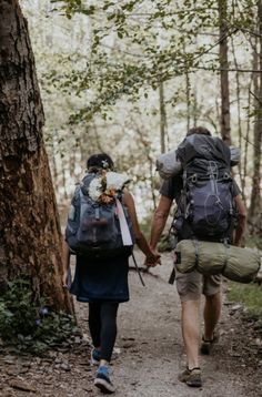 two people walking in the woods with backpacks on their back and one holding hands