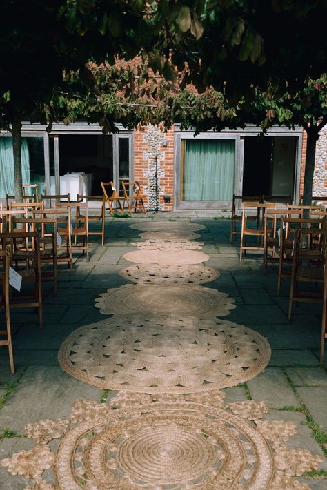 This shows an image showing a ceremony area at an outdoor wedding. On the aisle are different jute rugs lined to accentuate the aisle. Alongside the aisle are vintage folding chairs. Rugs Isle Wedding, Jute Rug Aisle Runner Wedding, Isle Rugs Wedding, Rugs For Outdoor Wedding, Round Rug Wedding Ceremony, Rugs At Wedding Ceremony, Rugs Aisle Wedding, Outdoor Wedding Rugs, Rug Isle Wedding