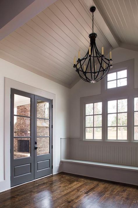 Gray cottage mudroom features a gray shiplap vaulted ceiling accented with an iron chandelier illuminating a gray beadboard window seat bench across from black glass paned doors. Beadboard Window, Gray Beadboard, Cottage Mudroom, Gray Cottage, Cottage Doors, Cottage Laundry Room, Gray Shiplap, Window Bench Seat, Farmhouse Mudroom