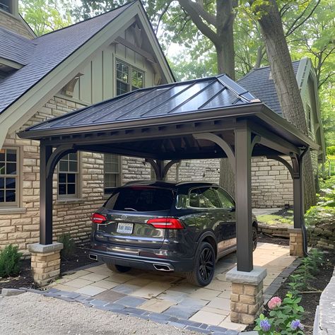 Elegant carport featuring a dark gray metal roof, beautifully complemented by tan stone accents and limestone piers. This open-sided structure shelters a black Volkswagen Tiguan, set against an old cottage-style house with black windows. The landscaping boasts lush greenery, colorful flowers, and two large decorative brown blocks enhancing the aesthetic. Carport Beside House, Beautiful Carport Ideas, Carport Driveway Ideas, Cottage Carport Ideas, Adding A Carport To Front Of House, Carport With Privacy Wall, Pergola As Carport, Covered Car Port Ideas, Pretty Carport Ideas