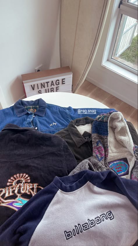 a stack of vintage surf brand and surf aesthetic jackets and jumpers with big 90’s surf branding lying on a table with a surfboard and a vintage surf sign in the background Vintage Surf Outfit Men, Vintage Billabong Jacket, Rip Curl Outfits, Surf Clothing Style, Vintage Surf Clothing, Vintage Surf Fashion, Rip Curl Aesthetic, Vintage Surf Aesthetic Outfit, 90s Surf Fashion