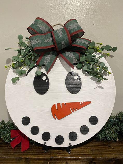 Welcome Winter and your guests with this 18" Snowman Door Hanger. Painted white and lightly weathered, with a green and red bow and a dusty colored greenery for a final touch! - Made From 1/4" Baltic Birch.- Measures 18" x 18" - Ready to hang! *Important Notes* We recommend all signs be hung indoors or in a covered outdoor area to keep your sign protected and beautiful! Processing Time And Shipping Due to demand each sign may take 1-2 weeks to cut, assemble, and package with lots of care. Please Wreath With Snowman, Round Snowman Door Hanger, Welcome Cricut Sign, Cricut Winter Door Sign, Christmas Painted Door Signs, Fall Front Door Signs Wooden, Christmas Door Hanging, Circular Christmas Sign, Easy Door Hangers Diy Crafts